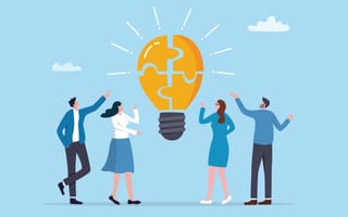An illustration of four professionals standing pointing toward a giant floating lightbulb composed of puzzle pieces, symbolizing the idea of teamwork