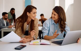 Two smiling women discuss a project together while working side by side in an office