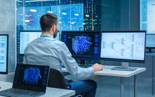 A man with a beard, seated at a workstation, analyzes a complex 3D data visualization on multiple monitors in a modern, high-tech server room.
