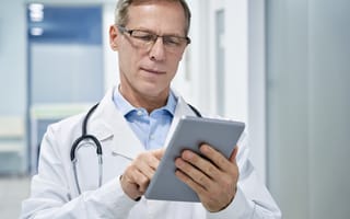 A focused doctor in a white coat and glasses uses a tablet in a hospital corridor.