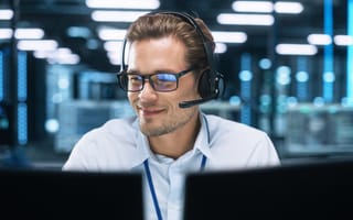 A man wearing glasses and a headset smiles while working on multiple monitors in a dimly lit tech environment, suggesting focus and expertise.