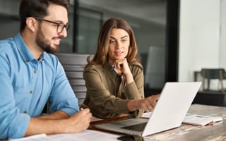 Two people at a desk, focused on a laptop. The woman in an olive blouse points at the screen, suggesting collaboration. The man in a blue shirt smiles.