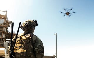 A soldier in a camouflage uniform controls a drone as it flies through the air.