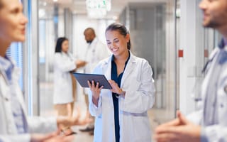A woman in a white coat holds a tablet device while surrounded by several other doctors in a healthcare setting.