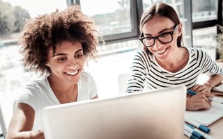 Two smiling women software engineers look at code on a laptop monitor while working together in an office