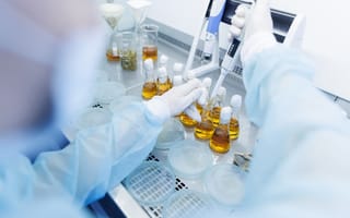 A pair of gloved hands dip a pipette into a test tube filled with yellow liquid to represent research