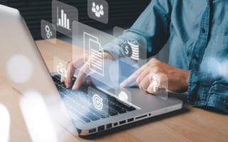 A person in a denim shirt types on a laptop, with semi-transparent digital icons — including graphs, documents, currency, and user profiles — floating above the keyboard, representing data analytics, finance, and business operations.