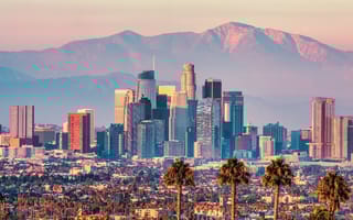 A view of the downtown Los Angeles skyline featuring palm trees and skyscrapers in the foreground and mountains in the background.
