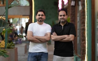 Two men stand confidently with arms crossed in a stylish setting. One wears a white shirt and jeans, the other a black shirt with light pants.