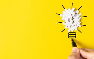 An image of a hand drawing a lightbulb on a bright yellow background with white puzzle pieces forming the top part of the lightbulb, symbolizing innovation