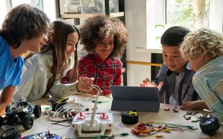 Five children gather around a tablet at a table with electronics and robotics, smiling and engaged.