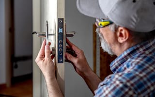 A man in a plaid shirt and cap works on a metal door lock with focus and precision.