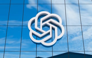 A white, abstract geometric logo on a reflective glass building facade, set against a blue sky with scattered clouds