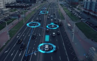 Aerial view of a busy city road with multiple cars. Blue digital overlays highlight vehicles, indicating autonomous driving technology. Modern and futuristic tone.