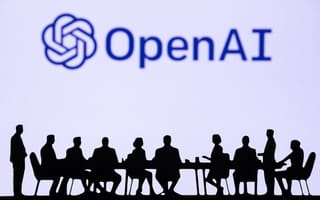 Several people sit around a table on top of a stage with the OpenAI logo in the background.