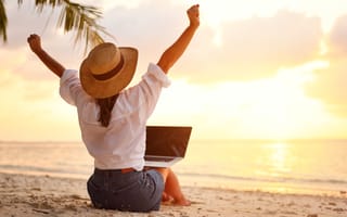 A woman stretches her arms above her head while working on her laptop on a beach at sunset