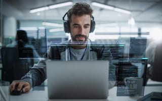 A focused man wearing headphones sits at a laptop in a modern office, surrounded by transparent screens displaying computer code, conveying a tech-savvy atmosphere.