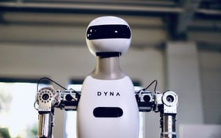 A humanoid robot with a sleek white exterior and black visor stands indoors. The word "DYNA" is on its chest, evoking a futuristic tone.