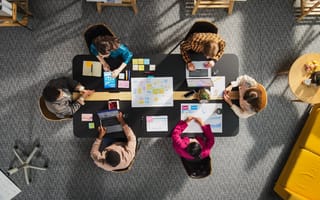 Overhead shot of five people collaborating around a black meeting table in an office. They are looking at laptops, tablets, and sticky notes on the table.