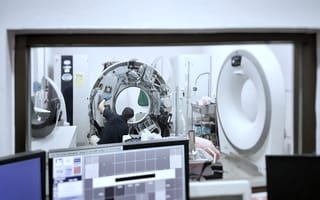 A technician repairs a disassembled MRI machine in a clinical setting, viewed through a window. Computer monitors in the foreground display data.