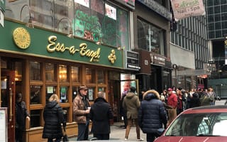 People stand in line outside "Ess-a-Bagel" on a busy city street. Signs for nearby salons appear above. Captures a bustling, chilly urban atmosphere.