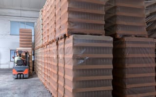 A forklift operator maneuvers between large stacks of boxes wrapped in brown plastic in a spacious warehouse.