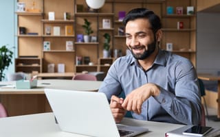 A man with a beard smiles while video conferencing on a laptop in an office. Books and plants are on shelves behind him. The mood is positive and focused.