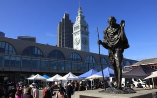 Statue of a robed figure holding a staff stands near tents at an outdoor market. Behind, a historic clock tower and skyscrapers under a clear blue sky.