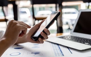 A person holds a smartphone, pointing at the screen. Nearby, an open laptop and graphs on paper suggest a business or office setting.