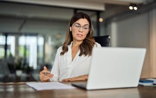 Businesswoman in glasses, wearing a white blouse, sits at a desk, focused on a laptop screen in a modern, well-lit office setting.