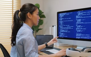 A woman in a blue shirt focuses intently on a desktop computer displaying lines of code. The background features a bright window with blinds and a plant, conveying a professional and concentrated atmosphere.