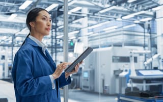 A woman in a blue lab coat holds a tablet, observing a modern, well-lit industrial facility with machinery.
