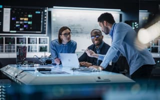 A diverse group of three professionals collaborates in a modern tech lab, engaging around a table with laptops and circuit boards, conveying teamwork and innovation.