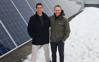 Two men stand next to solar panels in the snow wearing winter coats and boots and smiling at the camera.