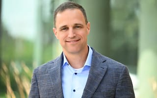 Matt Milton in a blue shirt and gray blazer stands outdoors, with a blurred green background.