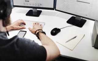 An employee wearing a blackshirt and a headset sits a desk typing on a keyboard in front of two desktop computer monitors.