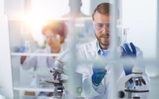 A scientist wearing a white lab coat, goggles and blue gloves is pictured sitting behind a microscope.