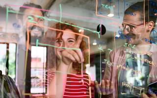 Four smiling software engineers outline a project on a translucent whiteboard while working together at a startup