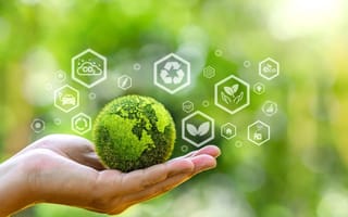A hand holds a globe covered in green grass and surrounded by icons that represent sustainability and renewable energy.