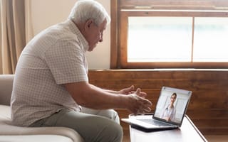 An elderly man sits on a couch engaged in a video call with a female doctor on a laptop. The room is softly lit with a large window behind him.