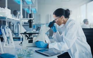 Hemab Therapeutics Raises $157M to Advance Bleeding Disorder Treatments A scientist in a lab coat and safety goggles examines a microscope in a bright laboratory. Blue gloves and lab equipment are visible, conveying focus.
