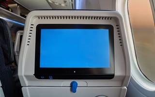 Platinum Equity Acquires Media Technology Provider Anuvu Airplane seat-back screen displaying a blank blue screen. Visible seat upholstery and aircraft window with a subtle view of the sky outside.