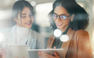 Two women smiling and engaged, holding digital tablets. The background is softly blurred with bright lights, conveying a modern, professional atmosphere.