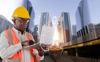 A construction worker in a yellow hard hat and orange vest uses a tablet with digital interface graphics overlaid. Modern skyscrapers rise in the background.