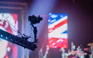 A camera on a crane faces a stage with vibrant lighting. A large, colorful LED screen in the background displays a Union Jack pattern.