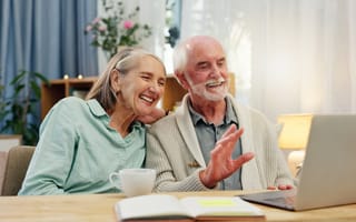 Elderly couple smiling while video chatting on a laptop in a cozy living room. They look joyful and relaxed, with plants and a warm lamp nearby.