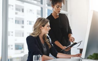 Two women in business attire work in a bright office. One is seated, focused on a computer, while the other stands, holding a notebook, conveying teamwork.
