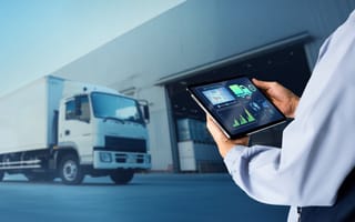 Person holding a tablet with logistics data charts, standing near a white delivery truck outside a warehouse. The scene conveys efficiency in transport management.