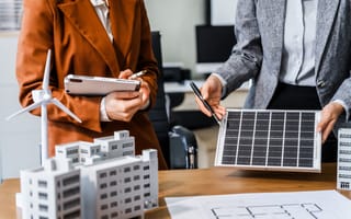 Two people in business attire discuss renewable energy. One holds a solar panel and a pen, while the other holds a tablet. Models of wind turbines and buildings are on the table.