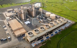 BKV Increases Stake in Joint Power Venture to Boost AI-Driven Energy Expansion Aerial view of an industrial power plant with cylindrical cooling towers, metal chimneys, and expansive green fields in the background under a clear sky.